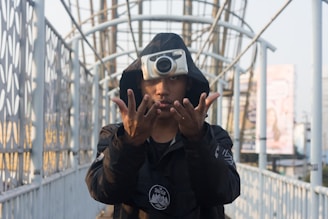 A person wearing a black hoodie is standing on a bridge. They are performing a trick, skillfully tossing a camera in mid-air, capturing the moment of suspension. The bridge structure is intricate with metal railings and background billboards.