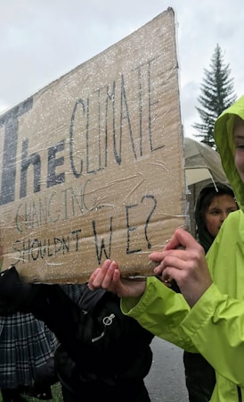 Several people are holding a cardboard sign covered in plastic. The text on the sign reads 'The climate is changing. Shouldn't we?' It appears to be raining, as there are water droplets visible.