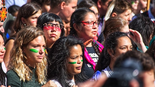A diverse group of people gathered closely together, many with colored face paint stripes, suggesting participation in an event or protest. The expressions range from serious to contemplative, indicating engagement or focus.