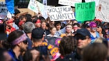 A large group of people engaged in a protest or rally. Many hold signs with various messages, including environmental and anti-capitalism statements. The crowd is diverse, with individuals wearing colorful clothing and face paint, conveying a sense of unity and activism. The atmosphere is lively and bustling.