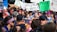 A large group of people engaged in a protest or rally. Many hold signs with various messages, including environmental and anti-capitalism statements. The crowd is diverse, with individuals wearing colorful clothing and face paint, conveying a sense of unity and activism. The atmosphere is lively and bustling.