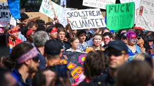 A large group of people engaged in a protest or rally. Many hold signs with various messages, including environmental and anti-capitalism statements. The crowd is diverse, with individuals wearing colorful clothing and face paint, conveying a sense of unity and activism. The atmosphere is lively and bustling.