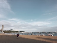A beach scene with a sandy shoreline and a view of the ocean populated with boats. In the distance, buildings are visible on the horizon, and the word 'Pattaya' is displayed on a hillside. The sky is mostly clear with some light clouds, and a few people are standing on the beach.