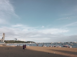 A beach scene with a sandy shoreline and a view of the ocean populated with boats. In the distance, buildings are visible on the horizon, and the word 'Pattaya' is displayed on a hillside. The sky is mostly clear with some light clouds, and a few people are standing on the beach.