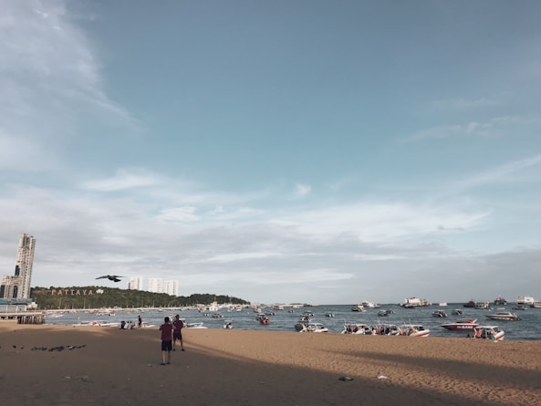 A beach scene with a sandy shoreline and a view of the ocean populated with boats. In the distance, buildings are visible on the horizon, and the word 'Pattaya' is displayed on a hillside. The sky is mostly clear with some light clouds, and a few people are standing on the beach.