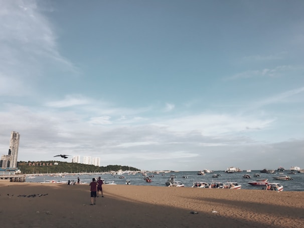 A beach scene with a sandy shoreline and a view of the ocean populated with boats. In the distance, buildings are visible on the horizon, and the word 'Pattaya' is displayed on a hillside. The sky is mostly clear with some light clouds, and a few people are standing on the beach.