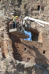 A pair of workers are on a boom lift near an ancient, weathered brick wall. They are wearing hard hats and safety vests, examining or repairing the historical structure.