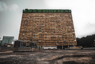 A large, rectangular apartment building stands prominently against an overcast sky. The structure features multiple rows of windows and air conditioning units, with a green top section. The foreground shows an expansive concrete surface leading up to the building, with some smaller buildings and vegetation visible in the background.