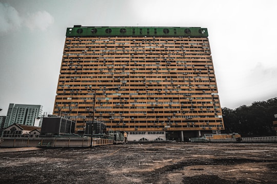 A large, rectangular apartment building stands prominently against an overcast sky. The structure features multiple rows of windows and air conditioning units, with a green top section. The foreground shows an expansive concrete surface leading up to the building, with some smaller buildings and vegetation visible in the background.
