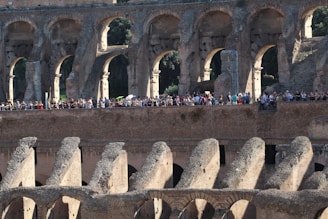An ancient amphitheater with multiple archways and rows of seating, bustling with tourists walking along the structure. The stone architecture is weathered yet majestic, highlighting the grandeur of the historic site.
