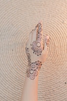 A hand adorned with intricate henna designs featuring floral patterns and swirls. The background is a textured, light-colored woven mat with concentric circles.