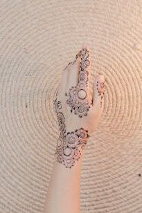 A hand adorned with intricate henna designs featuring floral patterns and swirls. The background is a textured, light-colored woven mat with concentric circles.