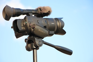 A professional Sony video camera mounted on a tripod. The camera has various controls and features, including a microphone with a windscreen, and is framed against a clear blue sky.