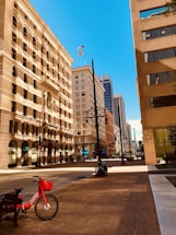 A quiet urban street is flanked by tall buildings with varied architectural styles under a clear blue sky. On the sidewalk, a red rental bike with the word 'JUMP' is parked near a bench. The wide street is empty, and flags are visible flying atop one of the buildings.