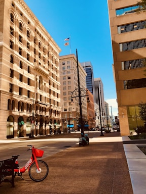 A quiet urban street is flanked by tall buildings with varied architectural styles under a clear blue sky. On the sidewalk, a red rental bike with the word 'JUMP' is parked near a bench. The wide street is empty, and flags are visible flying atop one of the buildings.