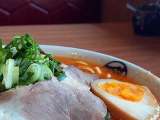 A close-up shot of a bowl of ramen featuring sliced pork, a halved soft-boiled egg with a rich yolk, and garnished with sliced green onions. The broth appears to be a vibrant orange, suggesting a spicy flavor. The bowl is set on a wooden table, with a blurred background hinting at a cozy restaurant setting.