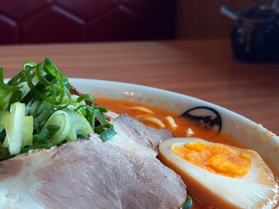 A close-up shot of a bowl of ramen featuring sliced pork, a halved soft-boiled egg with a rich yolk, and garnished with sliced green onions. The broth appears to be a vibrant orange, suggesting a spicy flavor. The bowl is set on a wooden table, with a blurred background hinting at a cozy restaurant setting.