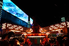 A brightly lit amusement park ride with an overhead structure adorned with numerous circular lights. The background features a large screen displaying a galactic or outer space scene with blue and purple hues. Neon lights and pillars enhance the futuristic ambiance, while a person walks past the ride.