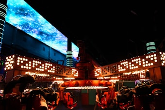 A brightly lit amusement park ride with an overhead structure adorned with numerous circular lights. The background features a large screen displaying a galactic or outer space scene with blue and purple hues. Neon lights and pillars enhance the futuristic ambiance, while a person walks past the ride.