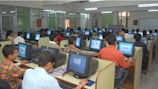 A large computer lab filled with many people seated at individual workstations. Each person is focused on the screen in front of them, with partitions separating each desk. The room is well-lit with fluorescent lights, and there are glass partitions creating smaller sections within the lab. The overall atmosphere suggests a busy, studious environment.