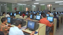 A large computer lab filled with many people seated at individual workstations. Each person is focused on the screen in front of them, with partitions separating each desk. The room is well-lit with fluorescent lights, and there are glass partitions creating smaller sections within the lab. The overall atmosphere suggests a busy, studious environment.