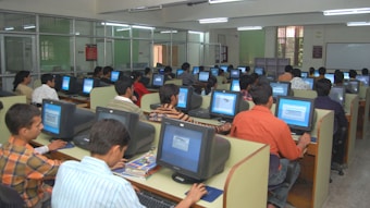 A large computer lab filled with many people seated at individual workstations. Each person is focused on the screen in front of them, with partitions separating each desk. The room is well-lit with fluorescent lights, and there are glass partitions creating smaller sections within the lab. The overall atmosphere suggests a busy, studious environment.