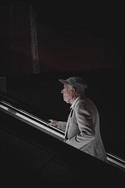 An elderly man in a light-colored suit and cap stands on an ascending escalator. His profile is illuminated by the light from the escalator, contrasting with the dark background.