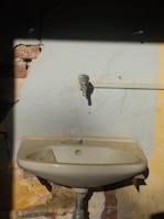 A damaged old sink is mounted on a wall with peeling paint and visible cracks. The wall shows signs of wear with some bricks exposed. There is a pipe running horizontally which appears disconnected from the faucet.