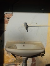 A damaged old sink is mounted on a wall with peeling paint and visible cracks. The wall shows signs of wear with some bricks exposed. There is a pipe running horizontally which appears disconnected from the faucet.