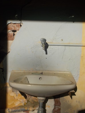 A damaged old sink is mounted on a wall with peeling paint and visible cracks. The wall shows signs of wear with some bricks exposed. There is a pipe running horizontally which appears disconnected from the faucet.