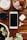 A wooden surface holds a smartphone, a pair of wireless earphones, a small black camera lens, and a potted plant. Nearby is a bowl filled with seashells and decorative objects, as well as a candle.