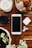 A wooden surface holds a smartphone, a pair of wireless earphones, a small black camera lens, and a potted plant. Nearby is a bowl filled with seashells and decorative objects, as well as a candle.