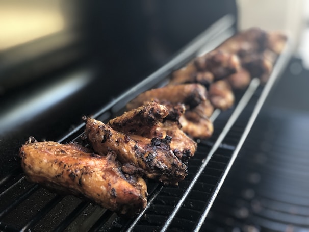 Grilled chicken wings are lined up on a barbecue grate, with a focus on the textured, charred surface of the meat. The background is slightly blurred, indicating depth of field, and enhancing the prominence of the cooked wings.