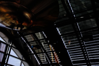 A large industrial ceiling with metal beams and grid-like structures. Light filters through slats, casting shadows and reflections. A dome-shaped metallic structure with perforations and warm lighting is prominently featured.