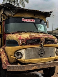 A rusty, old truck with a Mercedes-Benz emblem on the grille and weathered paint in shades of yellow and red. The windshield has a visible sticker reading 'Hyperlube Your Preferred Lubricant'. The truck is parked under a cloudy sky with palm trees and rooftops in the background. The condition indicates it is not in active use.