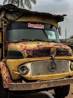 A rusty, old truck with a Mercedes-Benz emblem on the grille and weathered paint in shades of yellow and red. The windshield has a visible sticker reading 'Hyperlube Your Preferred Lubricant'. The truck is parked under a cloudy sky with palm trees and rooftops in the background. The condition indicates it is not in active use.