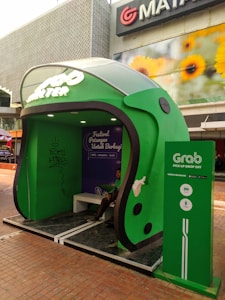 A large green structure designed to resemble a helmet functions as a waiting area or shelter. The branding indicates it is related to a ride-hailing service. A person is seated inside. The shelter is placed outdoors on a tiled surface with a building in the background displaying a sign with sunflowers.