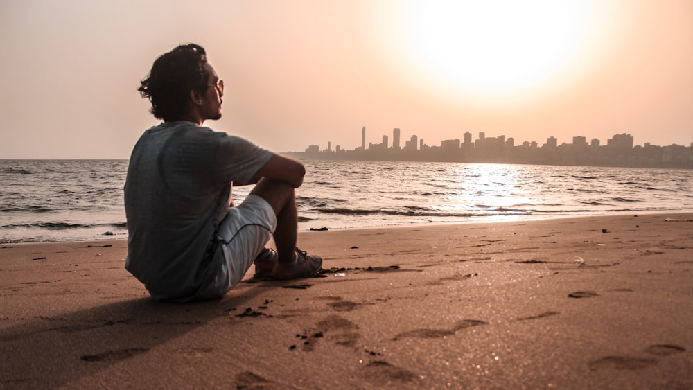 man in gray shirt sitting on sand near body of water