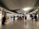 A spacious airport terminal corridor with a high curved ceiling and reflective tile flooring. Several shops line both sides, and numerous travelers are walking through or browsing. The lighting is bright, with decorative spotlights highlighting the shops.