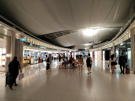 A spacious airport terminal corridor with a high curved ceiling and reflective tile flooring. Several shops line both sides, and numerous travelers are walking through or browsing. The lighting is bright, with decorative spotlights highlighting the shops.