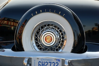 A close-up of the back of a vintage car showcasing a Cadillac emblem on a spare tire cover with intricate chrome detailing. The Ontario license plate is visible at the bottom.