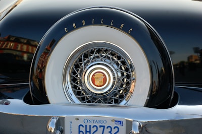 A close-up of the back of a vintage car showcasing a Cadillac emblem on a spare tire cover with intricate chrome detailing. The Ontario license plate is visible at the bottom.