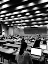 A large lecture hall filled with many students sitting at desks, focused on their laptops and notes. The room has a high ceiling with linear light fixtures, creating a structured atmosphere. The walls are lined with panels, and there is a large blackboard at the front of the room.