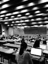 A large lecture hall filled with many students sitting at desks, focused on their laptops and notes. The room has a high ceiling with linear light fixtures, creating a structured atmosphere. The walls are lined with panels, and there is a large blackboard at the front of the room.