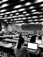 A large lecture hall filled with many students sitting at desks, focused on their laptops and notes. The room has a high ceiling with linear light fixtures, creating a structured atmosphere. The walls are lined with panels, and there is a large blackboard at the front of the room.