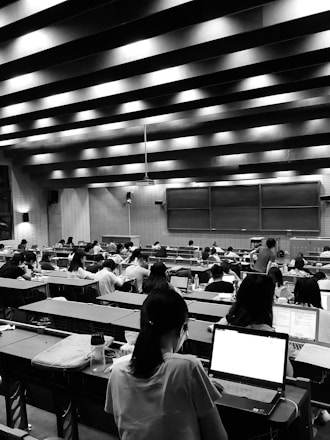 A large lecture hall filled with many students sitting at desks, focused on their laptops and notes. The room has a high ceiling with linear light fixtures, creating a structured atmosphere. The walls are lined with panels, and there is a large blackboard at the front of the room.