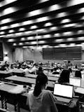 A large lecture hall filled with many students sitting at desks, focused on their laptops and notes. The room has a high ceiling with linear light fixtures, creating a structured atmosphere. The walls are lined with panels, and there is a large blackboard at the front of the room.