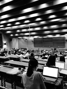A large lecture hall filled with many students sitting at desks, focused on their laptops and notes. The room has a high ceiling with linear light fixtures, creating a structured atmosphere. The walls are lined with panels, and there is a large blackboard at the front of the room.