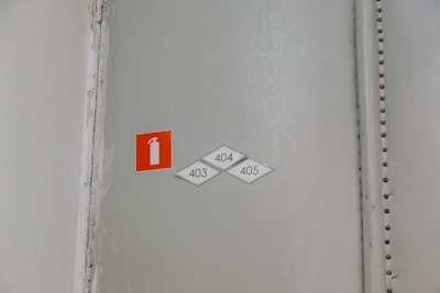 A wall with a small, rectangular red sign featuring a white fire extinguisher icon. Below the sign are three diamond-shaped labels with the numbers 403, 404, and 405. The wall is in a neutral color with a visible seam reinforced by metallic rivets.