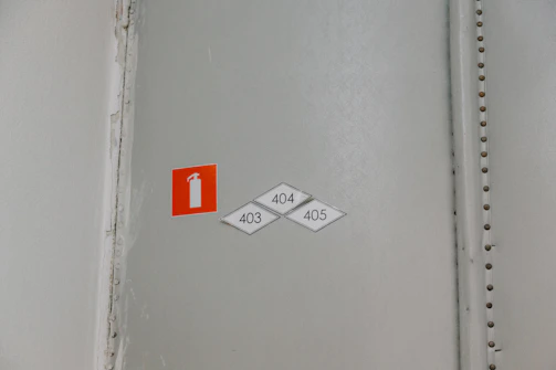 A wall with a small, rectangular red sign featuring a white fire extinguisher icon. Below the sign are three diamond-shaped labels with the numbers 403, 404, and 405. The wall is in a neutral color with a visible seam reinforced by metallic rivets.